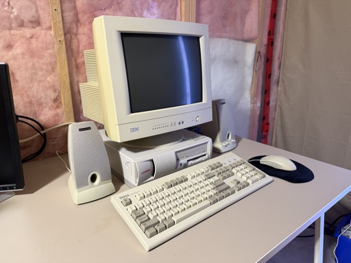 An old computer setup sits on a desk. A beige IBM CRT monitor is powered off, with a light reflection in the screen, and sits on top of a beige Compaq Deskpro EN. In front are a beige Dell keyboard and beige mouse on a mouse pad, with two beige Harman/Kardon speakers.