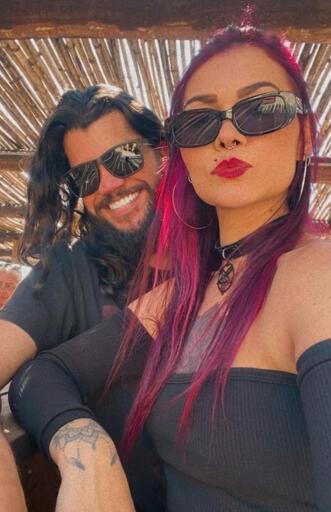 Two people are seated under a structure with horizontal wooden slats. The person on the left has long dark hair, a beard, and wears dark sunglasses and a black short - sleeve shirt. The person on the right has vibrant pink hair, dark sunglasses, red lipstick, a black choker with a pentagram pendant, an off - shoulder dark ribbed top, and visible tattoos on their arm. Both wear dark clothing. There is no text in the image.