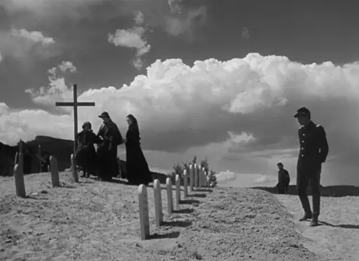 A cemetary on a hill, a soldier walks along a row of gravestones. In background at left , several mourners near a tall crucifix. Thick clouds loom in the disance.